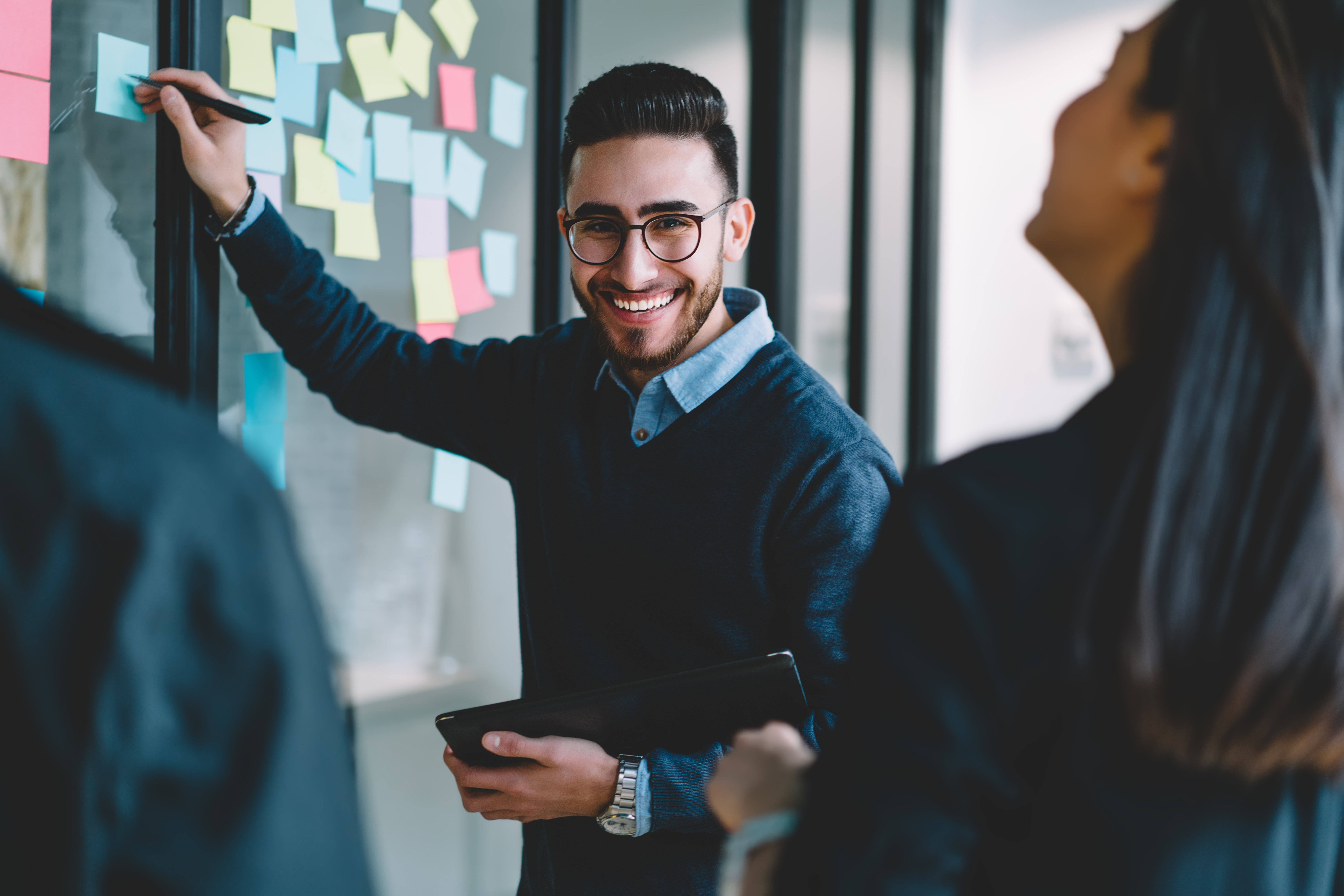 Half length portrait of happy young man smiling at camera while writing down notes on paper colorful stickers glued on wall during collaboration process with colleagues on studying courses in office